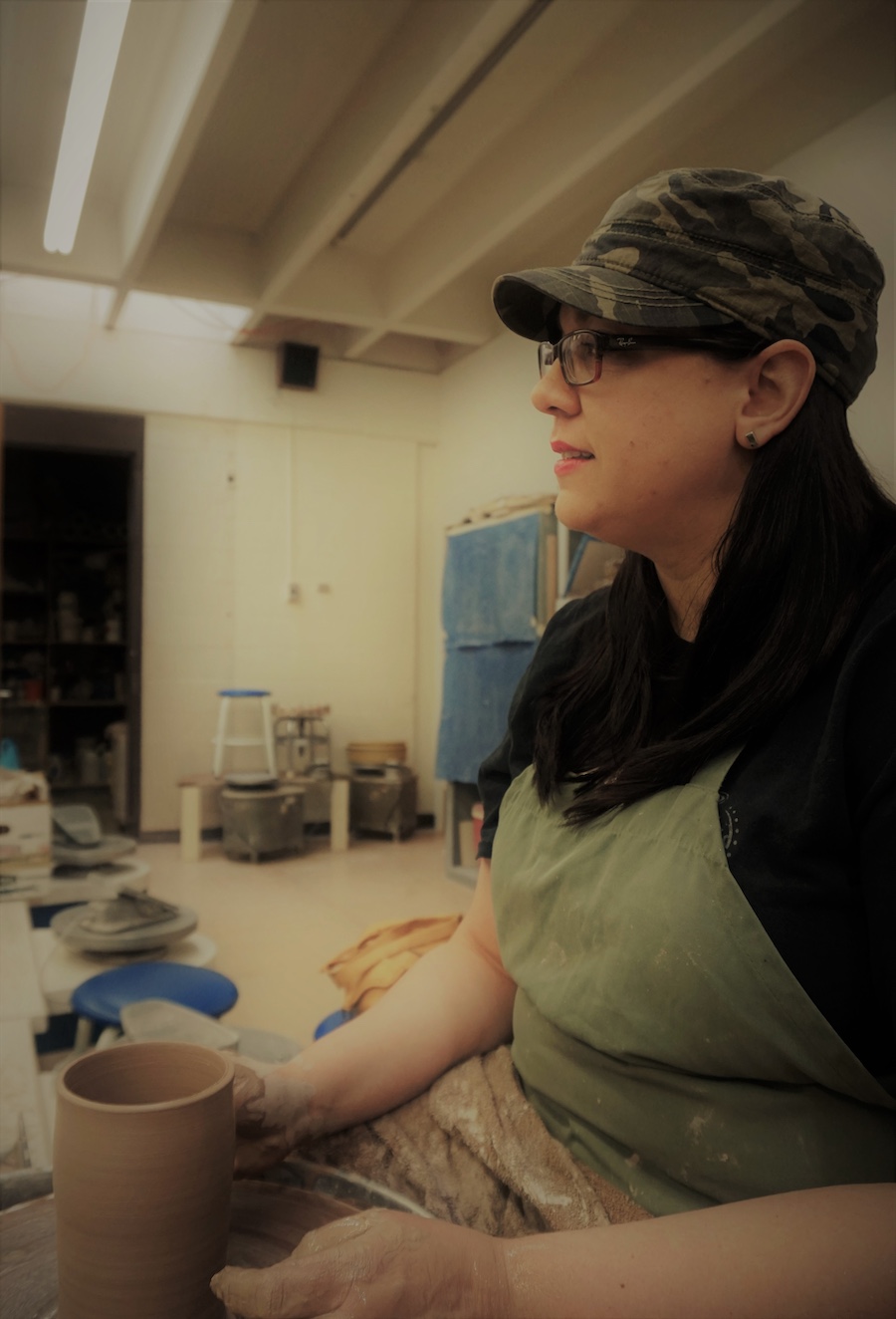 Sarah DeBaun wearing glasses, a camouflage cap, and an apron shapes clay on a pottery wheel in a ceramics studio, surrounded by shelves and pottery tools.