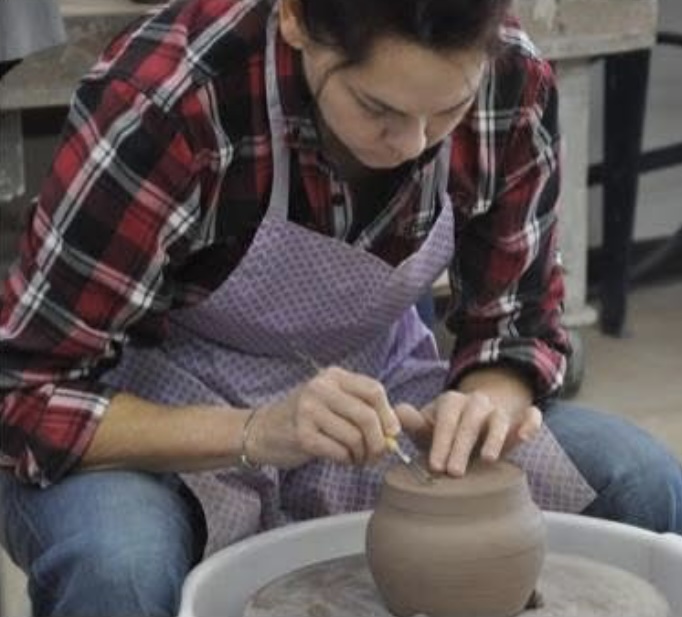 Screenshot Edie Hamilton wearing a plaid shirt and purple apron shapes a clay pot on a pottery wheel, carefully adding details with a small tool.