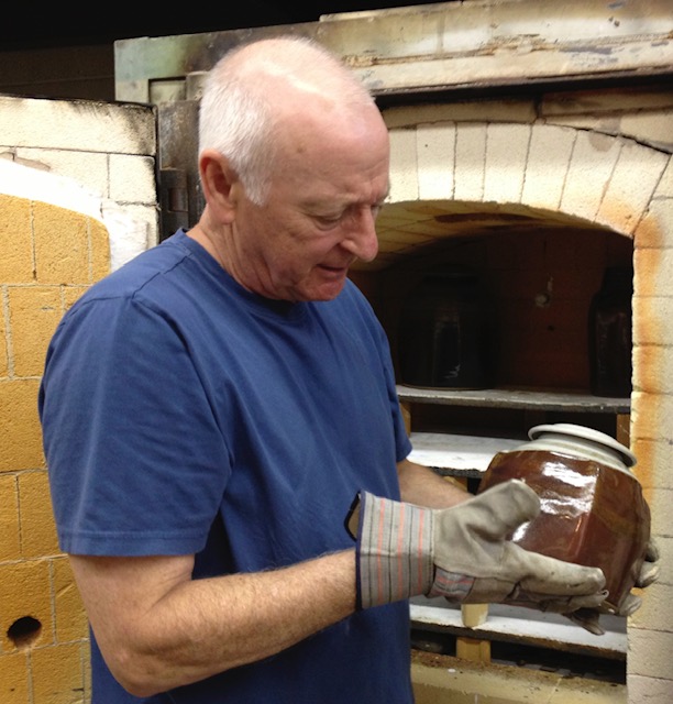 Frank Susi in a blue shirt and glove holds a ceramic pot, examining it closely in front of a kiln with an open door.