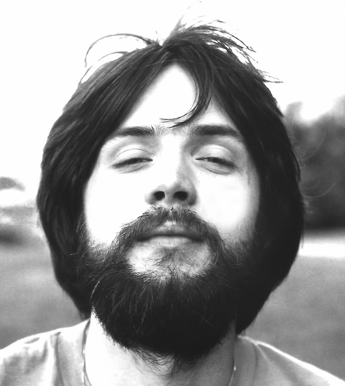 A young man with medium-length dark hair and a full beard is outdoors, eyes partially closed, and facing the camera with a relaxed expression. The background is blurred greenery.