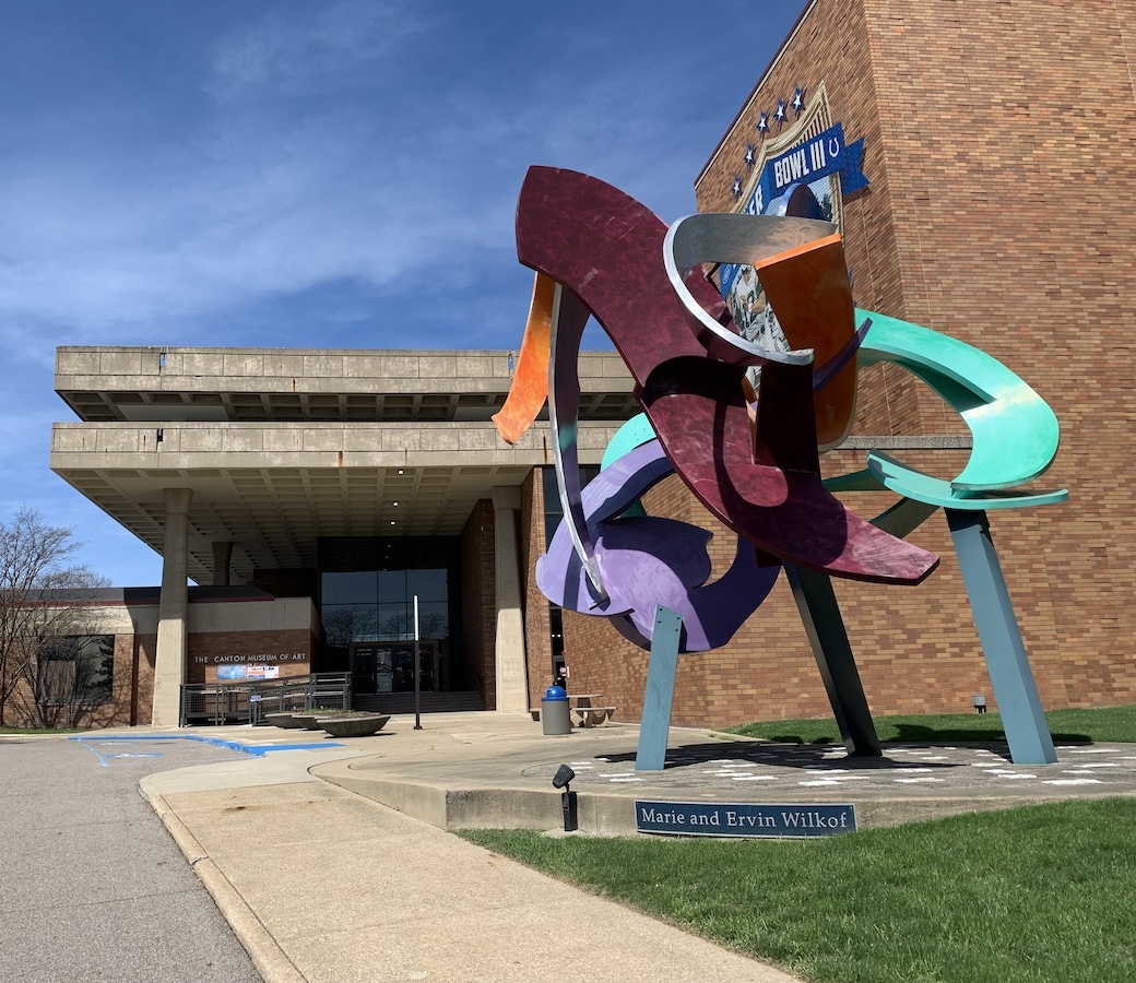CMA Colorful abstract metal sculpture stands in front of a brick building with a glass entrance. A sign reads “Marie and Ervin Wilkof.” Blue sky and some trees are visible in the background.